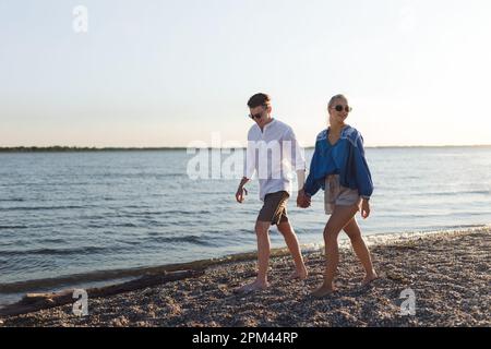 Glückliches junges Paar, das bei Sonnenuntergang am Strand spaziert und sich auf ein Picknick vorbereitet. Stockfoto