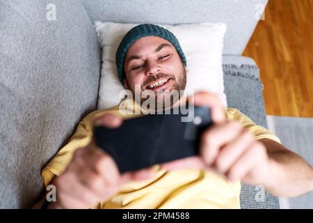 Mann, der sein Mobiltelefon für soziale Medien nutzt, während er sich zu Hause auf dem Sofa entspannt. Stockfoto