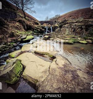 Three Shire Heads, wo das Wasser über moosbedeckte Felsen stürzt und eine Packpferdbrücke über den Bach ragt. Grüne, sanfte Hügel des Embras des Peak District Stockfoto