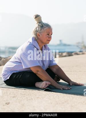Eine ernsthafte Frau im Alter mit Dreadlocks-Frisur, die am Strand gegen das Meer steht und an sonnigen Tagen die Pose oder Asana trainiert Stockfoto
