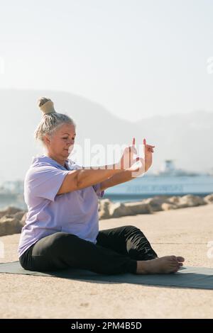 Eine ernsthafte Frau im Alter mit Dreadlocks-Frisur, die am Strand gegen das Meer steht und an sonnigen Tagen die Pose oder Asana trainiert Stockfoto