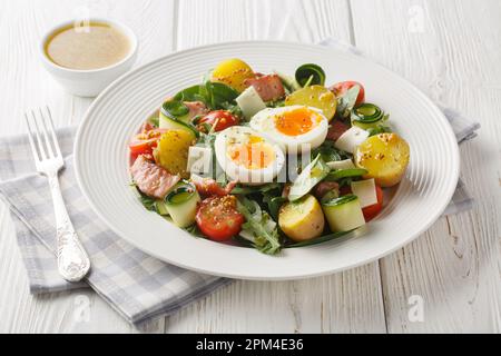 Bauernsalat mit Kartoffeln, Tomaten, Speck, Rucola, Zucchini, Käse und weich gekochte Eier, gewürzt mit Vinaigrette-Sauce, Nahaufnahme auf einem Teller auf dem T Stockfoto