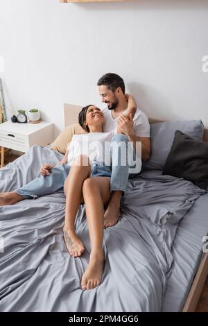Fröhliches, multiethnisches Paar, das sich zu Hause im Bett entspannt und sich anlächelt, Stockbild Stockfoto