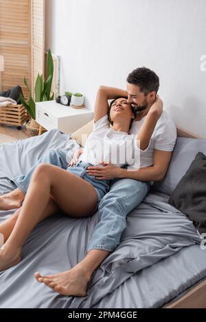 Junge afroamerikanische Frau in Jeansshorts umarmt einen bärtigen Mann, während sie sich zu Hause im Bett entspannt, Stockbild Stockfoto