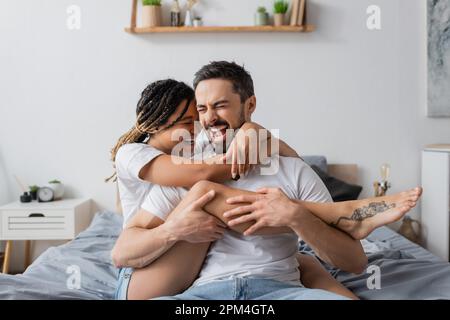 Aufgeregtes gemischtrassiges Paar in weißen T-Shirts, das sich umarmt, während es zu Hause im Schlafzimmer Spaß hat, Stock Image Stockfoto