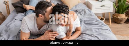 Fröhlicher Mann, der jungen afroamerikanischen Frau ein Geheimnis erzählt, während er sich zu Hause im Bett entspannt, Banner, Stock Image Stockfoto