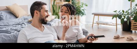 Aufgeregter bärtiger Mann fütterte afroamerikanische Frau mit Popcorn, während sie zu Hause im Schlafzimmer fernsah, Banner, Stock Image Stockfoto