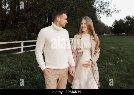 Ein junges, glückliches, romantisches schwangeres Paar geht an warmen Herbsttagen mit Händchen nach draußen. Schwangere Frau in Strickkleid und ihr gutaussehender Mann erwarten Stockfoto