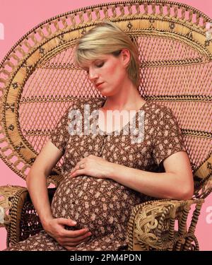 Studioporträt einer jungen, schwangeren Frau, die im Rattanstuhl sitzt. Stockfoto