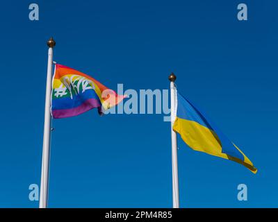 Die ukrainische Flagge und die Regenbogenflagge auf den Flaggenmasten am Hintergrund eines Himmels. Zwei Flaggen und ein wolkenloser Himmel. Stockfoto