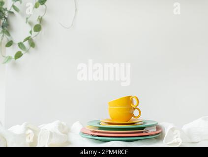 Leere gelbe Tassen und farbenfrohe Teller auf einem weißen Tisch, der mit einer Leinentischdecke bedeckt ist, und einen Blumentopf an der Wand im Hintergrund. Preparatio Stockfoto