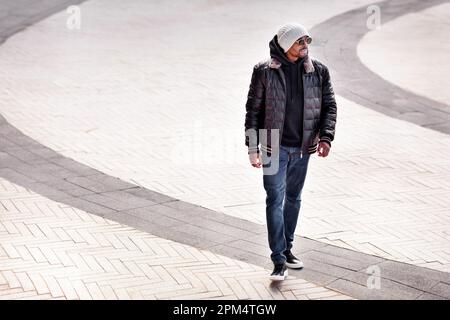 Street Fashion, stylischer afroamerikanischer Mann mit schwarzer Lederjacke, Hoodie, Sneakers, Jeans, Beanie und Sonnenbrille, der auf grauem Straßenrücken läuft Stockfoto