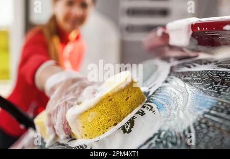 Junge Frau putzt die Heckscheibe ihres Autos, Nahaufnahme in Handschuhen mit gelbem Schwamm Stockfoto