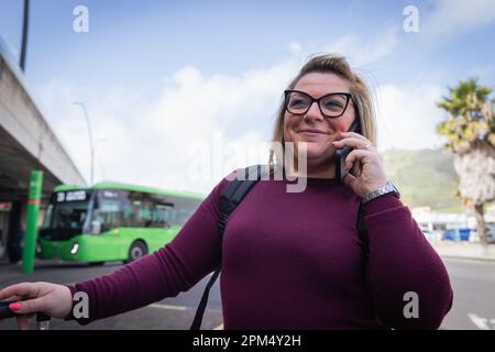 Eine lächelnde junge Frau telefoniert, während sie am Bahnhof auf den Bus wartet Stockfoto