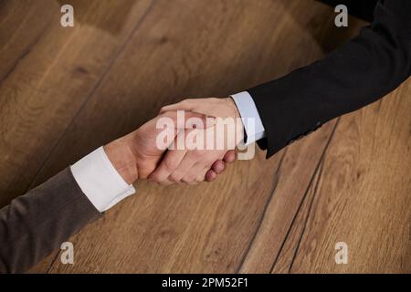 Nahaufnahme der Hände von zwei jungen und erfolgreichen männlichen Geschäftsleuten, die sich die Hände über einen Holztisch schütteln Stockfoto