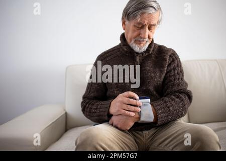 Seniormann, der ein medizinisches Gerät zur Blutdruckmessung verwendet - Älterer Mann, der an Bluthochdruck leidet, sitzt zu Hause auf dem Sofa und kümmert sich um seine h Stockfoto