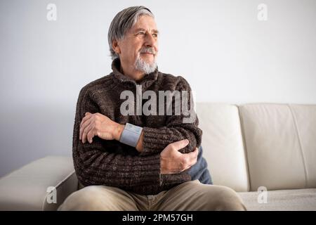 Seniormann, der ein medizinisches Gerät zur Blutdruckmessung verwendet - Älterer Mann, der an Bluthochdruck leidet, sitzt zu Hause auf dem Sofa und kümmert sich um seine h Stockfoto