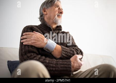 Seniormann, der ein medizinisches Gerät zur Blutdruckmessung verwendet - Älterer Mann, der an Bluthochdruck leidet, sitzt zu Hause auf dem Sofa und kümmert sich um seine h Stockfoto