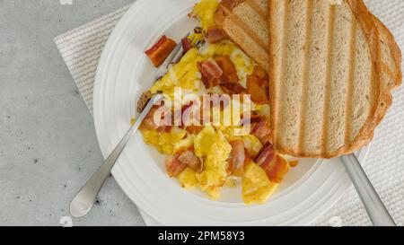 Englisches Frühstück. Rührei, gebratener Speck und Käse, serviert mit Toast. Essen auf einem Teller, flach liegend Stockfoto