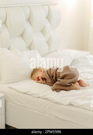 Ein Junge im braunen Bademantel liegt auf einem weißen Bett und schaut durch seine Hände. Vertikaler Rahmen. Stockfoto