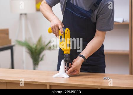 Erfahrener Zimmermann in blauen Overalls, mit gelbem Elektrobohrschrauber, Möbelmontage. Stockfoto