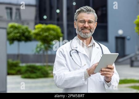 Porträt eines älteren grauhaarigen Arztes, der ausserhalb der Klinik steht, in einem weißen Kittel mit Stethoskop. Er hält ein Tablet in der Hand, sieht in die Kamera und lächelt. Stockfoto