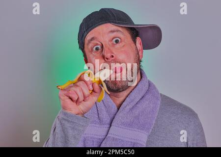 Bärtiger Mann mit Baseballkappe und Handtuch um den Hals, der eine Banane isst. Modellfreigabe verfügbar. Stockfoto