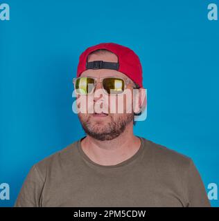 Bärtiger Mann mit Sonnenbrille und Mütze vor blauem Hintergrund. Modellversion verfügbar. Stockfoto