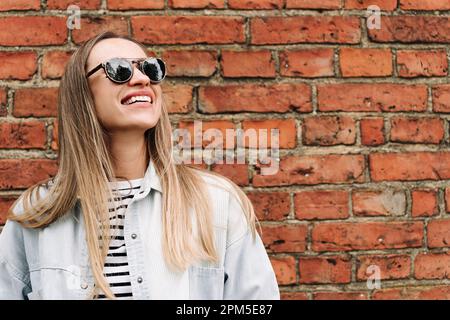 Eine langhaarige Frau mit Sonnenbrille lacht an der Ziegelwand Stockfoto