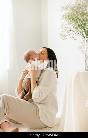 Sitzende junge Mutter küsst den neugeborenen Sohn am Fenster und Blumen Stockfoto