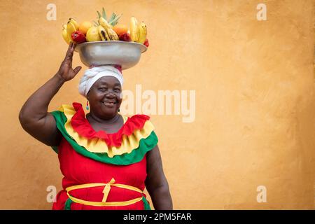 Fröhlicher, lächelnder Palenquera-Straßenverkäufer mit frischem Obst in der Altstadt von Cartagena, Kolumbien. Fröhliche afrokolumbianische Frau in traditionellen Kostümen. Stockfoto