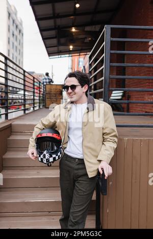 Ein glücklicher Mann, der einen Motorradhelm hält, der steht und lächelt Stockfoto