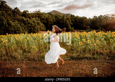 Ein junges Mädchen in fließendem weißen Kleid, das auf dem Feld der Sonnenblumen tanzt Stockfoto