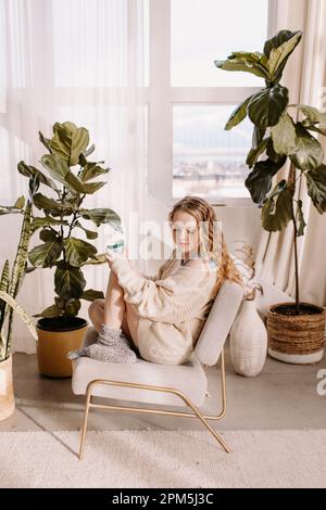 Gemütliche junge Frau in übergroßem Pullover, Füße zum Stuhl hochgezogen Stockfoto
