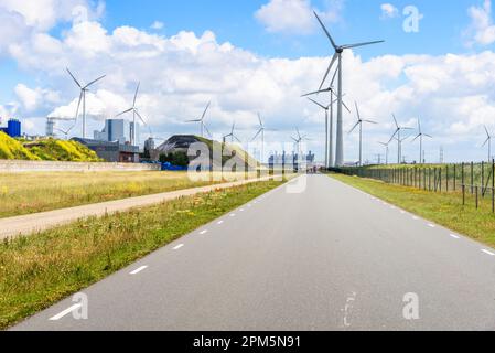 Die von Windturbinen gesäumte Straße führt an einem sonnigen Sommertag durch einen Industriepark. Auf der linken Seite des Bildes ist ein oal-befeuertes Kraftwerk zu sehen. Stockfoto