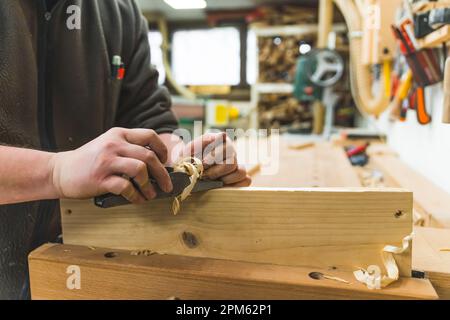 Männlicher Zimmermann, der mit einem Flugzeug auf einer Holzbohle arbeitet. Holzbearbeitungs-Werkstattinnenraum im Hintergrund. Hochwertiges Foto Stockfoto