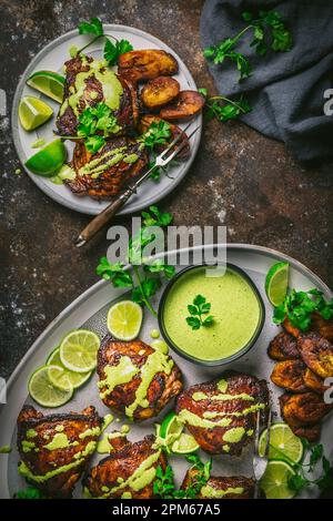 Peruanisches Hähnchenschenkel auf Platte und Servierteller, mit gebratenen Kochbananen, grünem Nieselregen und Limonen- und Kräutergarnierung Stockfoto