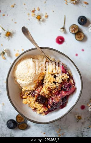 Apfel und Heidelbeere zerfallen in einer Schüssel mit einer Portion Vanilleeis. Ein alter Löffel ist in die Schüssel eingekeilt. Stockfoto