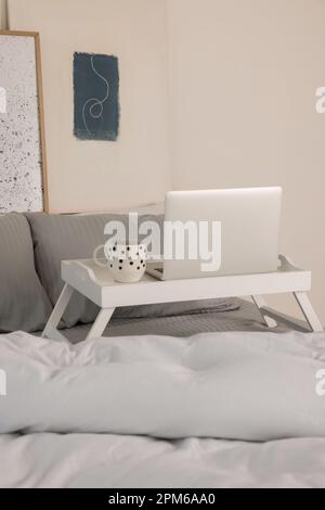 Weißer Tablett-Tisch mit Laptop und eine Tasse Getränk auf dem Bett drinnen Stockfoto