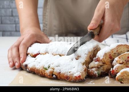 Eine Frau schneidet traditionelle Weihnachtsstollen mit Puderzucker auf einem weißen Holztisch, Nahaufnahme Stockfoto