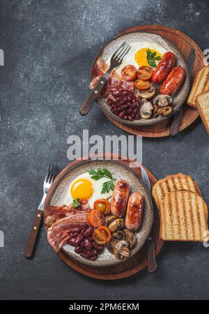 Traditionelles englisches Frühstück mit Spiegeleiern, Würstchen, Bohnen, Pilzen, gegrillten Tomaten, Speck und Toast auf grauen Tellern Stockfoto