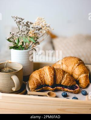 Frühstück im Bett mit frisch gebackenen Croissants mit Blaubeermarmelade und Tee in einer Tasse. Stockfoto