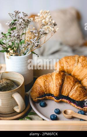 Frühstück im Bett mit frisch gebackenen Croissants mit Blaubeermarmelade und Tee in einer Tasse. Stockfoto