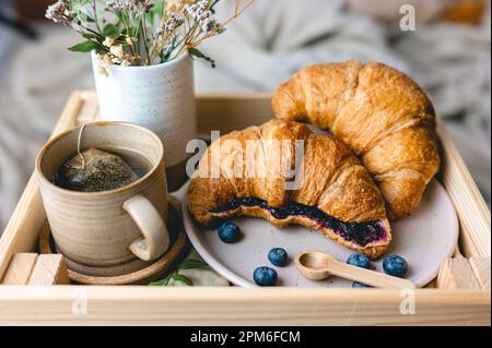 Frühstück im Bett mit frisch gebackenen Croissants mit Blaubeermarmelade und Tee in einer Tasse. Stockfoto