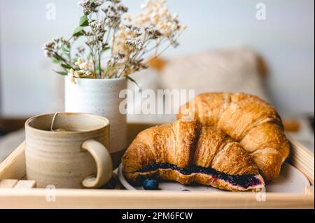 Frühstück im Bett mit frisch gebackenen Croissants mit Blaubeermarmelade und Tee in einer Tasse. Stockfoto
