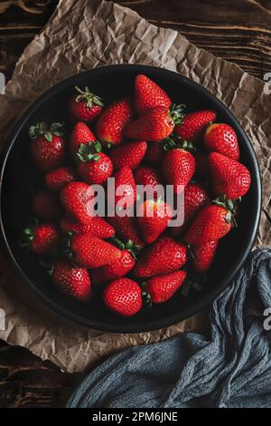 Schwarze Schüssel voller frischer Erdbeeren auf zerknittertem Papier, mit grüner Serviette, über einem Holztisch Stockfoto