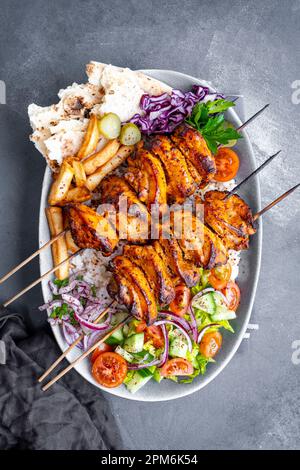 Hausgemachtes Hühnerdoner-Kebab auf Holzspießen, serviert auf Reis, garniert mit Pommes Frites, Gurken, Salat und Lavaschbrot auf einem ovalen Teller. Stockfoto