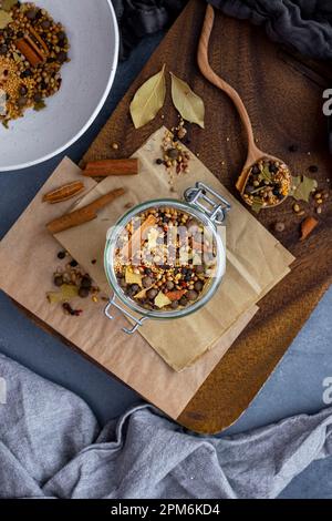 Senfkörner, Koriandersamen, Gewürznelken, Zimtstangen, Lorbeerblätter und rote Pfefferflocken, vermischt in einem Glasgefäß und einem Holzlöffel an der Seite. Stockfoto