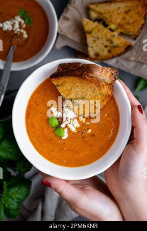 Frauenhände halten eine Schüssel Tomatensuppe, garniert mit Fetakäse und Basilikumblättern, und ein gegrilltes Käsesandwich, das darin getaucht wurde. Stockfoto