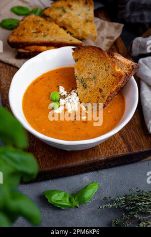 Geröstete Tomatensuppe mit Fetakäse und Basilikumblättern in einer weißen Schüssel, gegrilltes Käse-Sandwich und mehr gegrilltes Käse-Sandwich Stockfoto